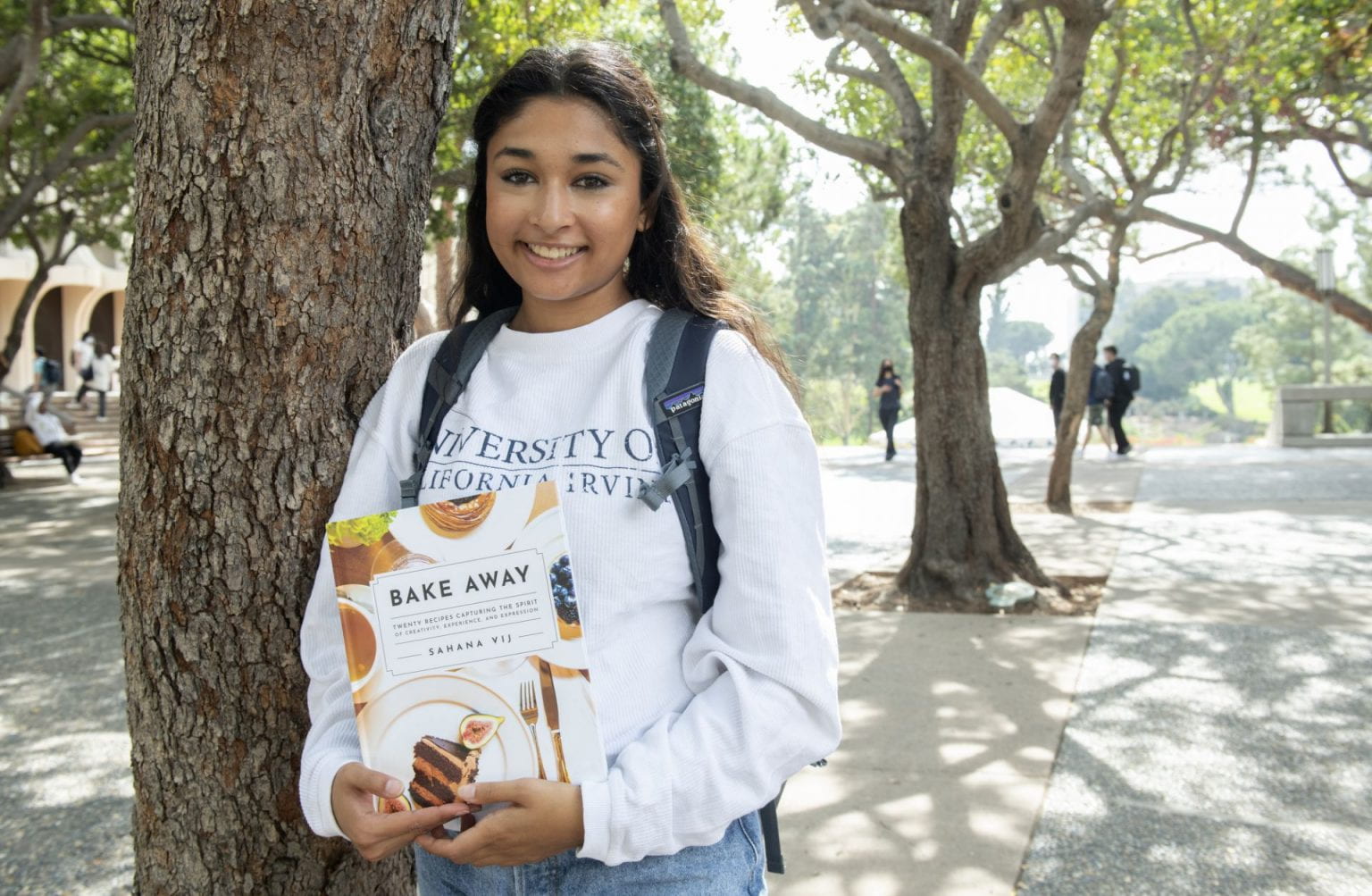 UICI student Sahana Vij standing in front of a tree and holding the cookbook Bake away that she wrote.