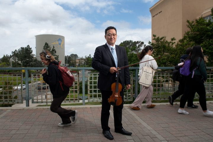 Dennis Kim, holding his violin and bow stands on a bridge with students in the background.