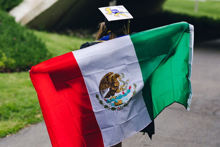 Jessica Ortiz wearing a cap and gown, while holding a Mexican flag across her back as she walks away from the camera.