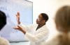 A man with googles and a white lab coat facing a white board with graphs.