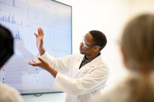 A man with googles and a white lab coat facing a white board with graphs.