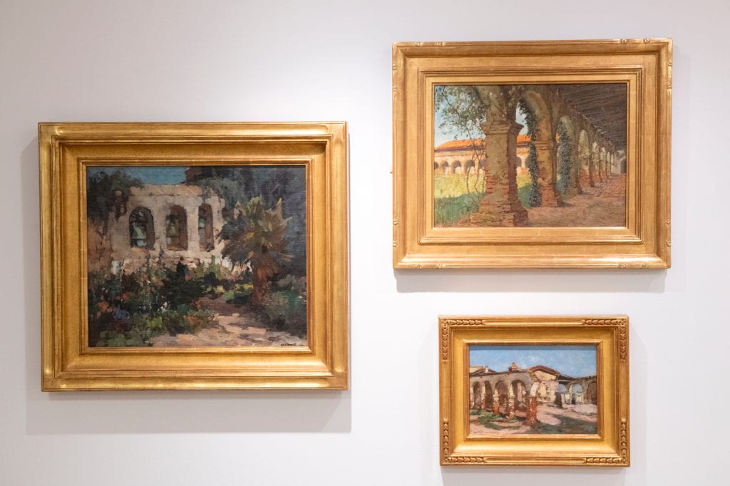 One section of the exhibition focuses on paintings of California missions, including (clockwise from left) George K. Brandriff’s “The Bells of Old San Juan” (Oil on canvas, 19 x 23 in.), William Adam’s “Back Corridor, San Juan Capistrano Mission” (1894, Oil on board, 16 x 21 in.) and George Gardner Symons’ “The Arches” (Oil on canvas, 10 x 14 in.)