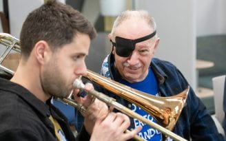 Rupert Whitehead, a trombonist with the Royal Philharmonic Orchestra, plays next to Terry Taylor, who had a stroke three years ago and whose right eye was affected by Bell’s palsy after a COVID-19 shot. Taylor, who also has spells of vertigo, wears a shirt that says, “I’m not clumsy. I’m performing random gravity checks.”